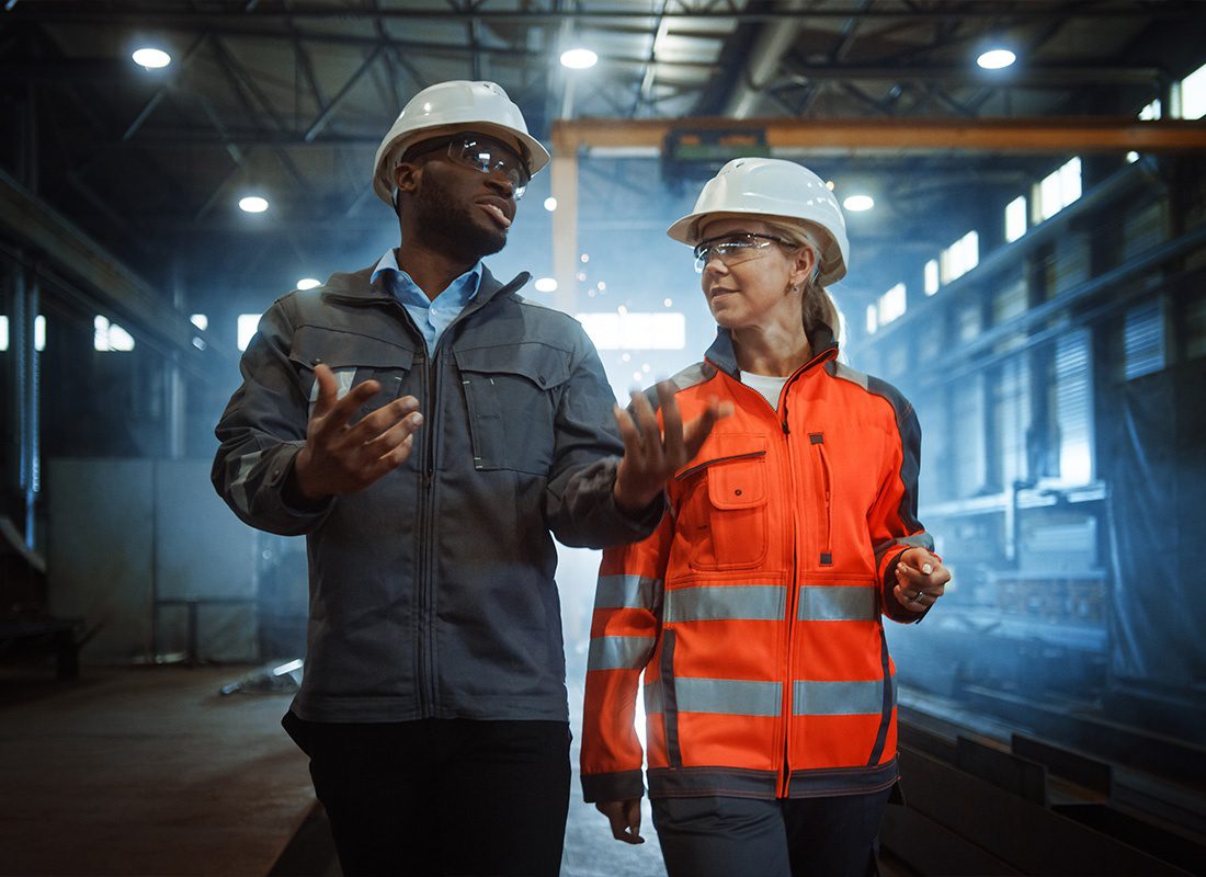Insurance by Industry - Two Heavy Industry Engineers in Hard Hats Walking in a Steel Metal Manufacturing Factory and Having a Discussion